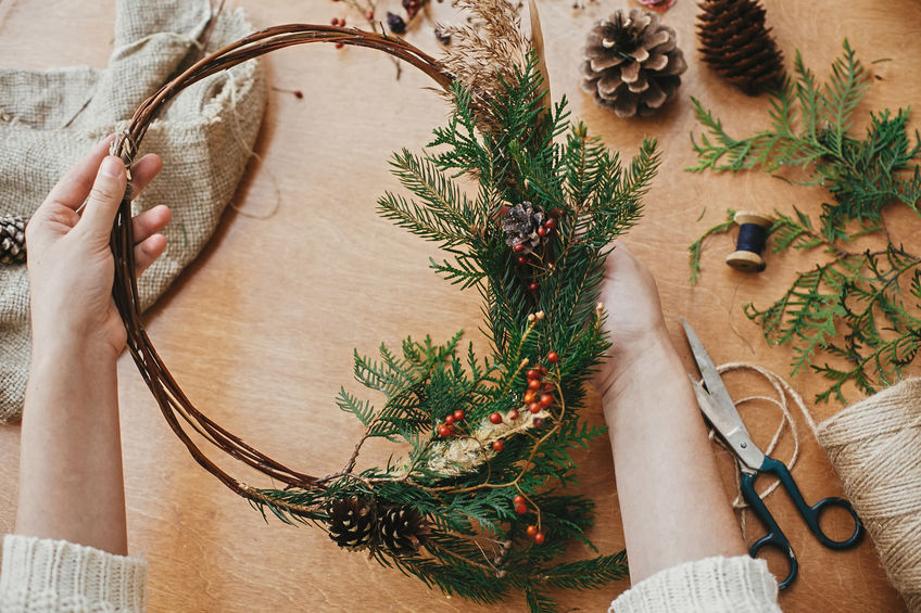 Hands holding rustic christmas wreath with pine cones, berries, fir branches, thread, scissors on wooden table.  Authentic rural wreath. Christmas wreath workshop.